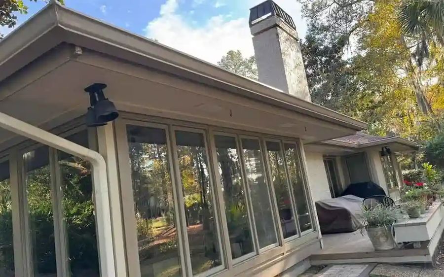 Almond seamless gutter and round downspout along the eave of a screened porch, with a chimney and trees behind
