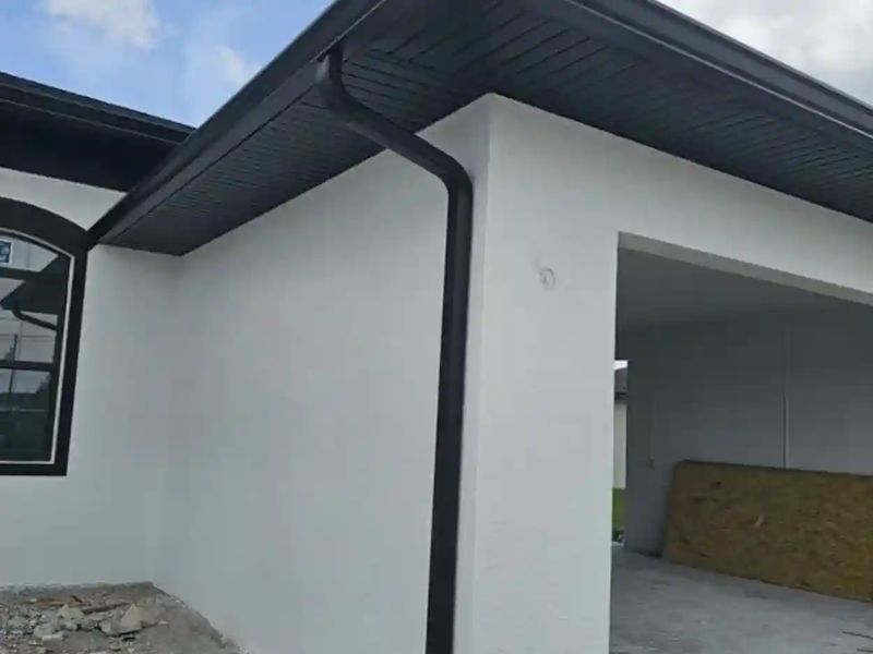 Black seamless gutter and downspout under a dark soffit on a white stucco house with a carport