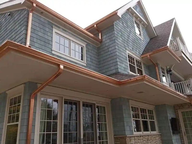 Copper gutters and round copper downspouts along the eaves and porch roof of a two-story house with blue-gray shingle siding