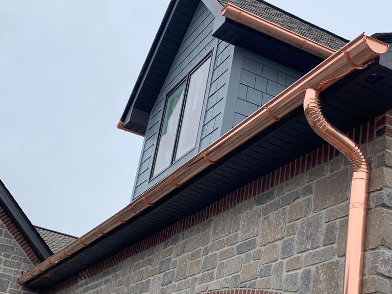 A copper half-round gutter with a round copper downspout on a house with stone veneer and dark siding under a dormer