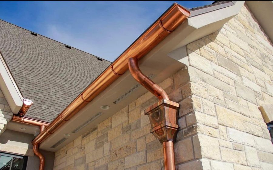 Copper K-style gutter with a copper leader head and downspout on a stone-clad house wall