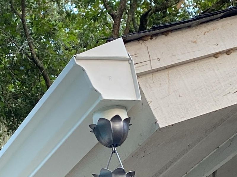 A cup-style metal rain chain hanging from the outlet of a white K-style gutter, with trees behind