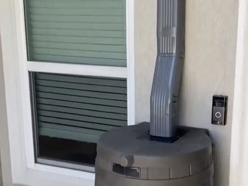 A gray downspout emptying into a brown rain barrel beside a window and front-door camera