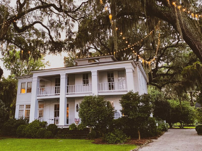 A white two-story Southern home with double porches under live oaks strung with lights