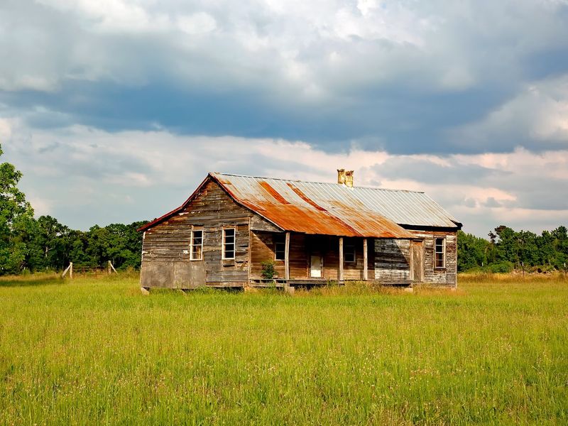 An old unpainted wooden farmhouse with a rusted tin roof and a deep front porch, standing in a grassy field
