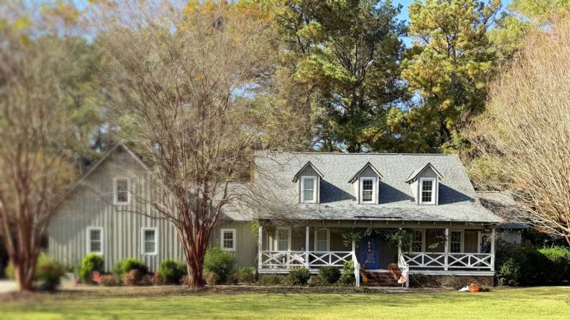 Board-and-batten Southern house with three dormers, a white-railed front porch, crape myrtles and tall pines behind it