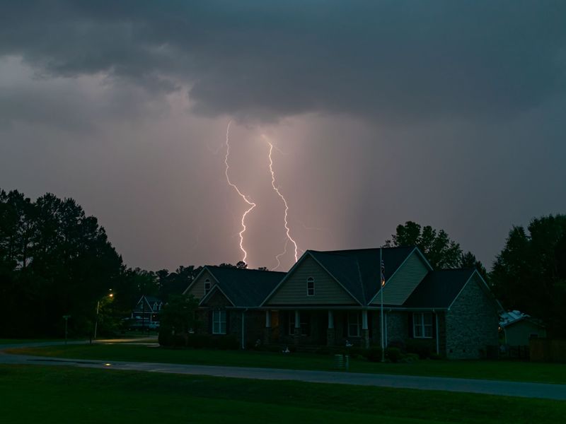Lightning forking over a brick and siding house among tall pines on a stormy evening