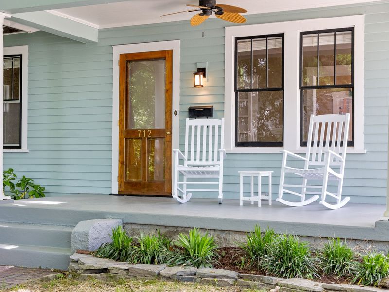 A front porch with two white rocking chairs, a wooden front door and pale blue-green siding