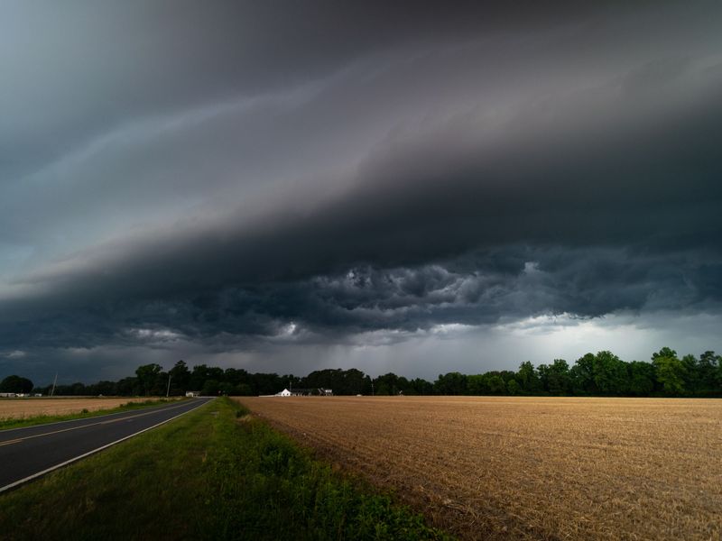 A dark shelf cloud rolling over a two-lane country road, a harvested field and a distant tree line