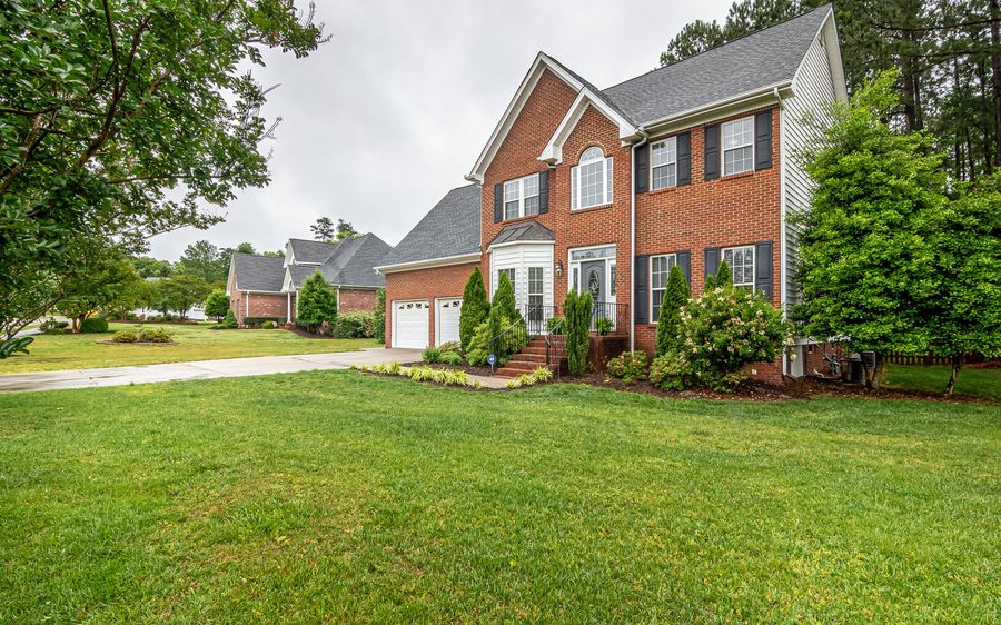 A two-story red brick house with white gutters, black shutters and a pine-lined lawn on an overcast day