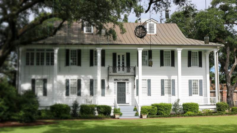 A white two-story Southern home with dark shutters, a metal roof and live oaks shading a wide front lawn