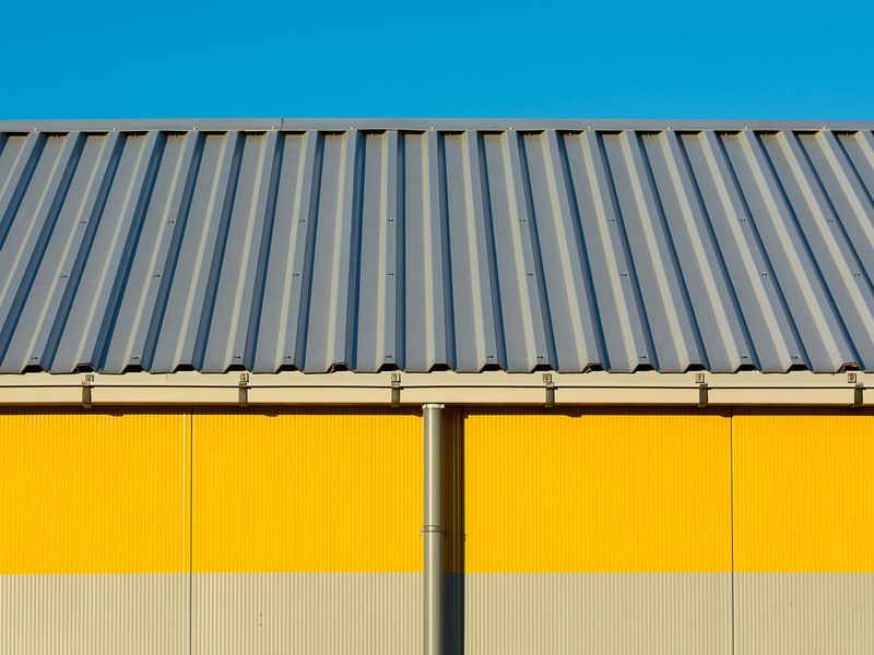 Yellow warehouse wall under a ribbed metal roof, with a gutter along the eave and a downspout