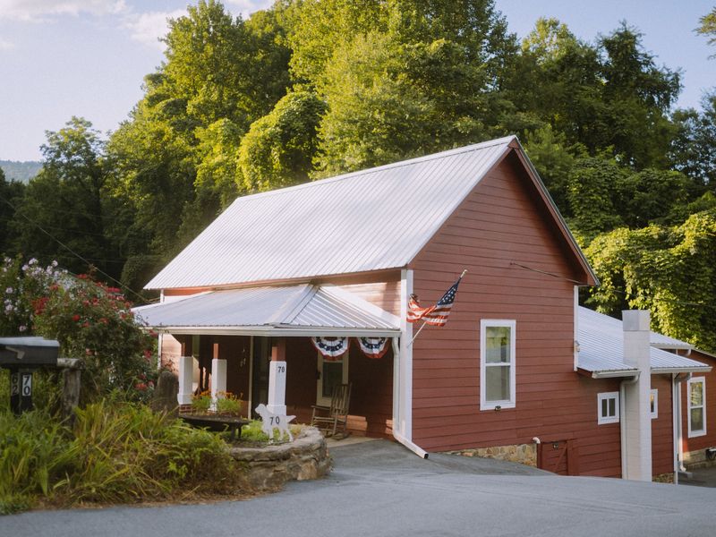 Red wood-sided house with a white metal roof, a covered front porch and a downspout at the corner, set against green trees