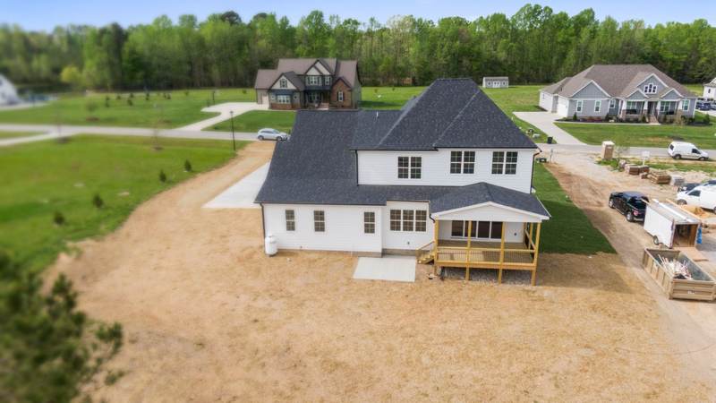 A newly built two-story house on a flat, freshly graded lot, its gutters not yet hung, with finished homes and a tree line beyond