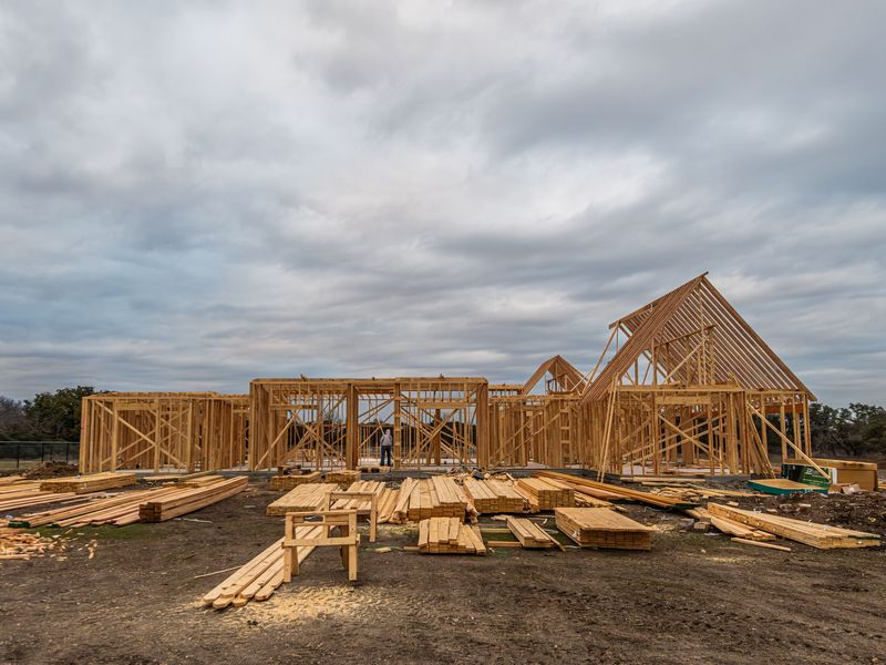 Wood framing and roof trusses for a new house on a cleared lot, with stacks of lumber in front under a cloudy sky