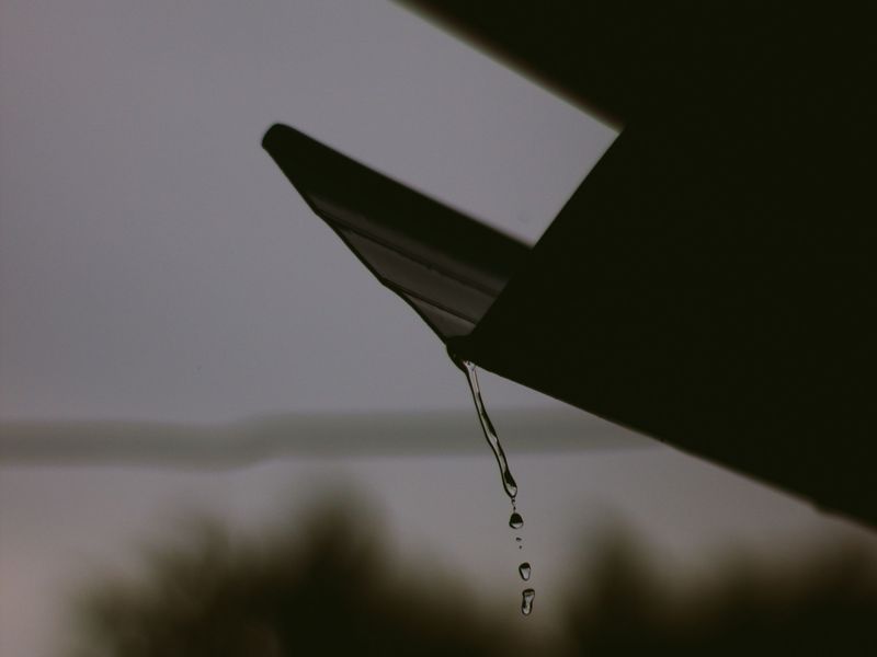 Rainwater dripping off the corner of a roof edge against a grey sky