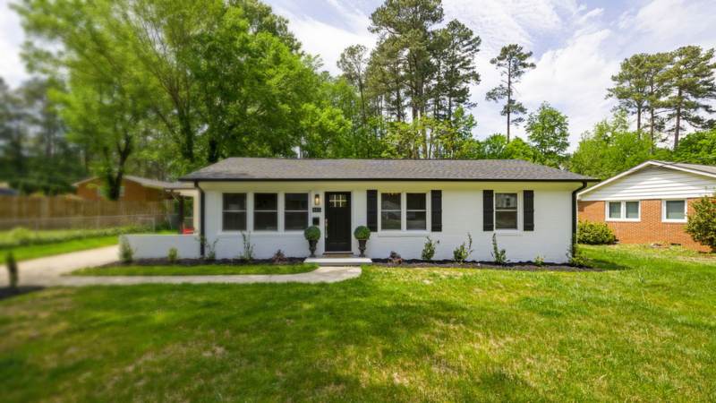Single-story painted brick ranch house with gutters and downspouts under tall pines