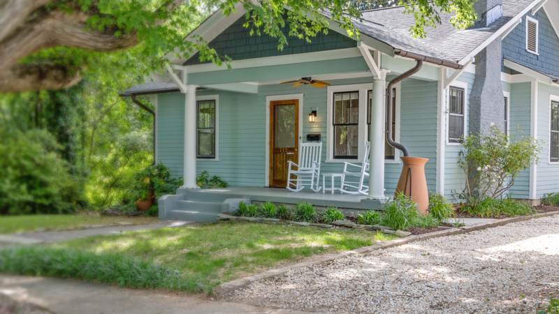 A pale blue bungalow with a front porch, white rocking chairs and a downspout emptying into a terracotta urn
