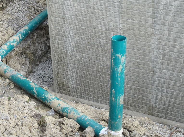A PVC drain line laid in a gravel-lined trench beside a house foundation, with a vertical clean-out riser