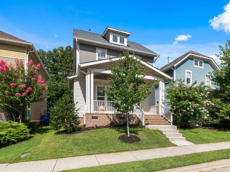 A gray craftsman-style house with a front porch on brick piers, beside a pink crape myrtle in bloom