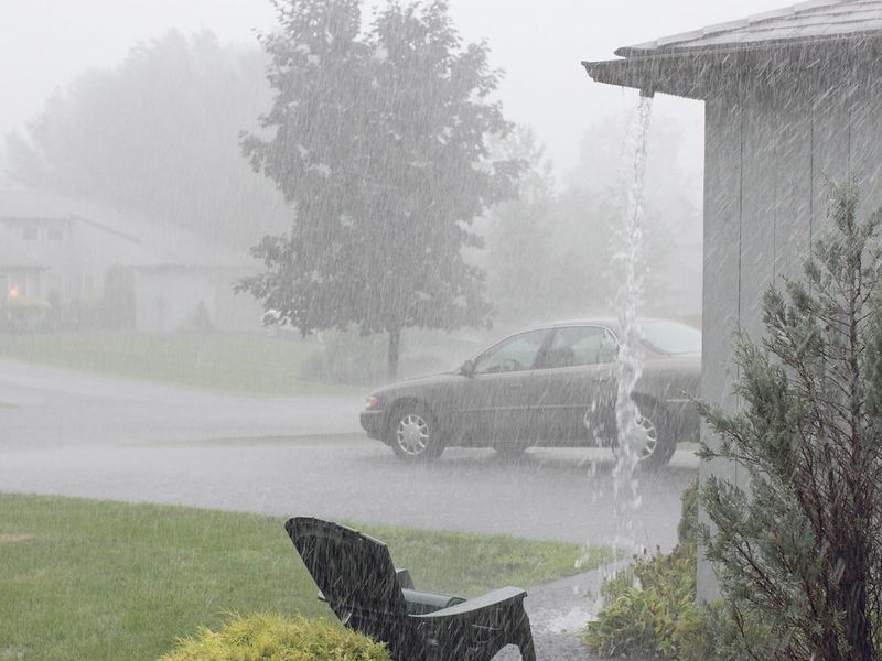 Rain pouring off the corner of a roof with no downspout during a heavy downpour on a suburban street