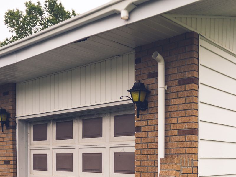 A white gutter and downspout on the brick corner of an older house, above a faded garage door