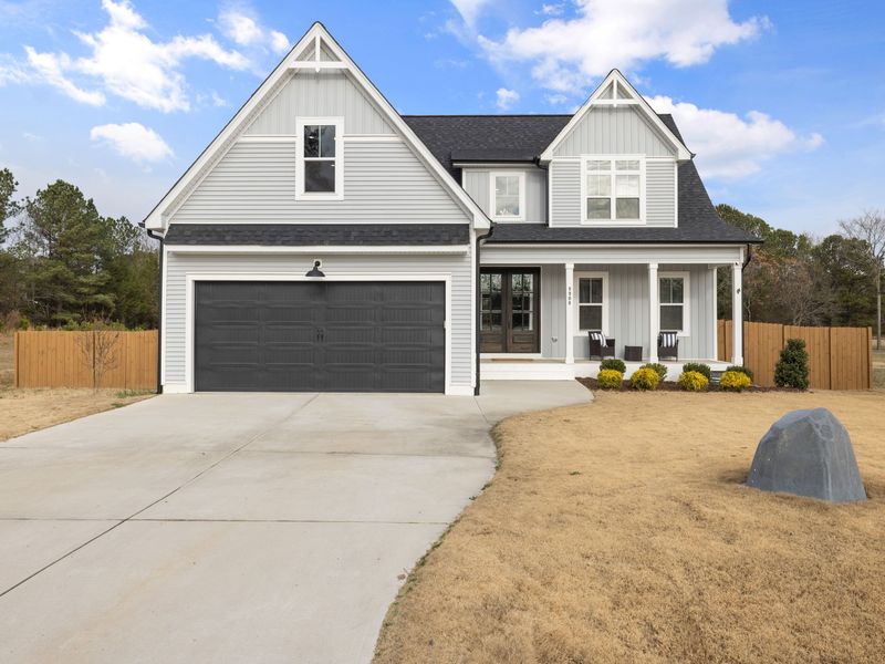 A gray two-story house with a front porch and a black garage door, with pine trees behind it