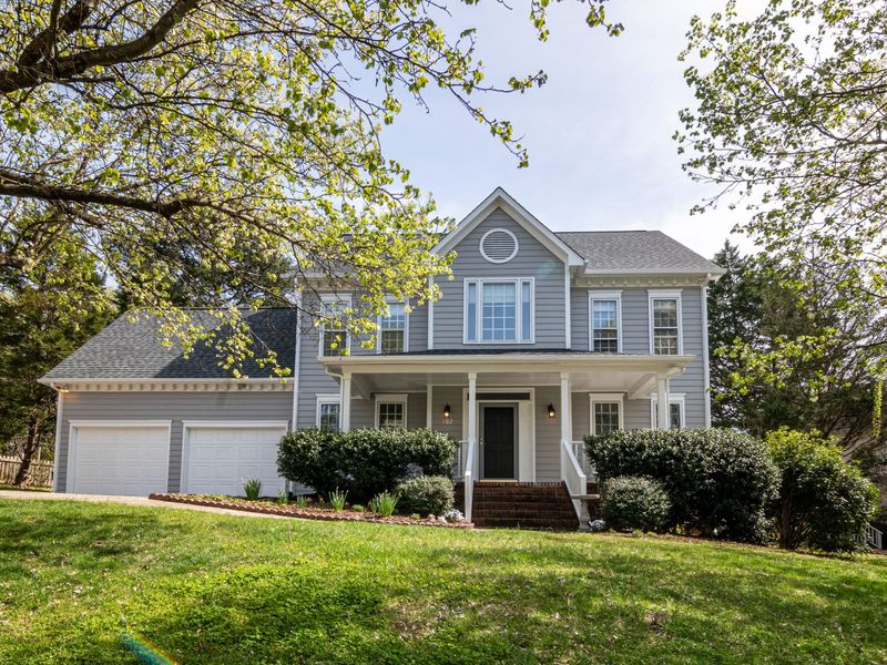 A gray two-story house with a front porch and a two-car garage, framed by spring tree branches above a sloping lawn