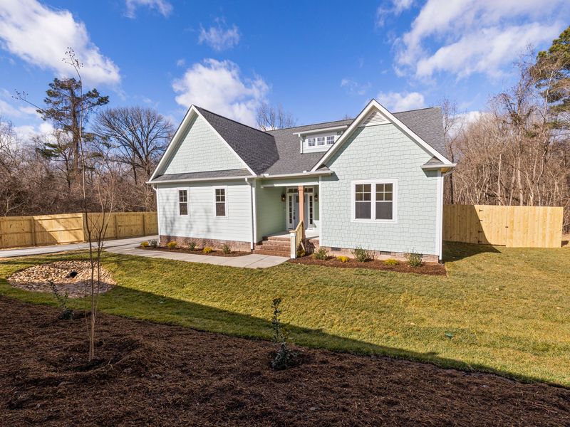 A newly built one-story house with pale green siding and bare white fascia, no gutters yet, on a fresh lawn backed by pines and bare hardwoods