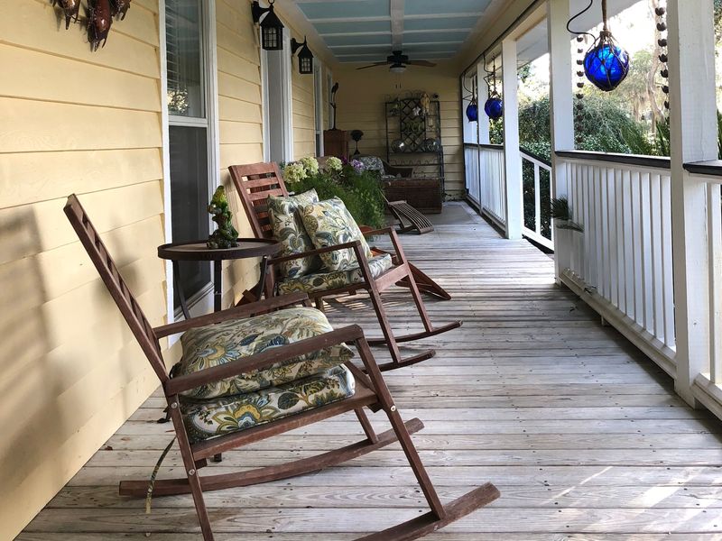 A long covered porch with wooden rocking chairs, ceiling fans and a pale blue ceiling, looking out toward moss-hung trees
