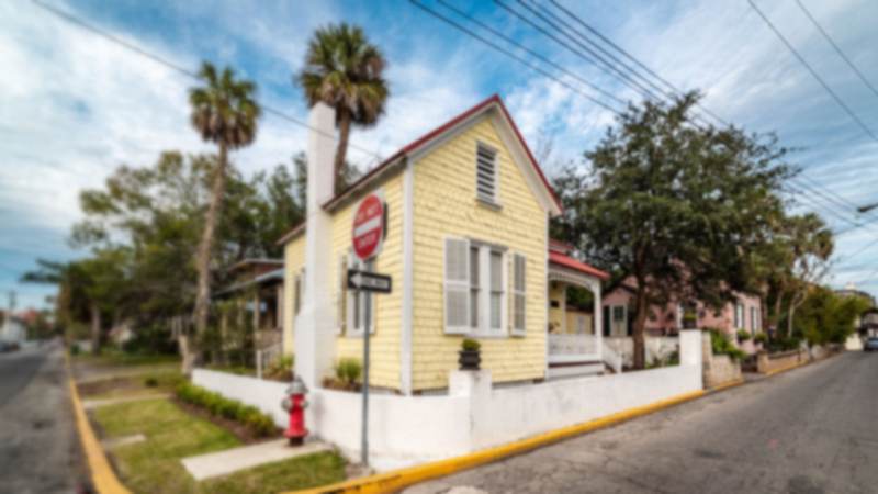Yellow shingle-sided cottage with a red metal roof and a white porch on a street corner in St. Augustine, with palm trees and neighboring historic homes