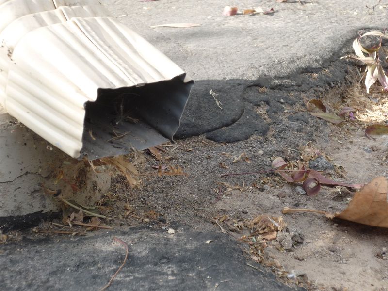 A dented downspout outlet emptying straight onto bare dirt beside a paved edge, with a dark wet patch below it