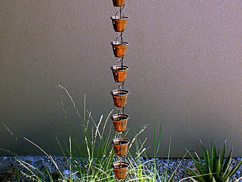 A copper cup rain chain hanging against a stucco wall into a gravel bed planted with grasses