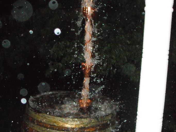 A copper rain chain streaming with water in a heavy night rain, splashing into a wooden barrel beside a white porch post