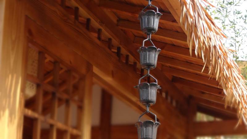 A cup-style rain chain hanging from the timber eave of a garden pavilion