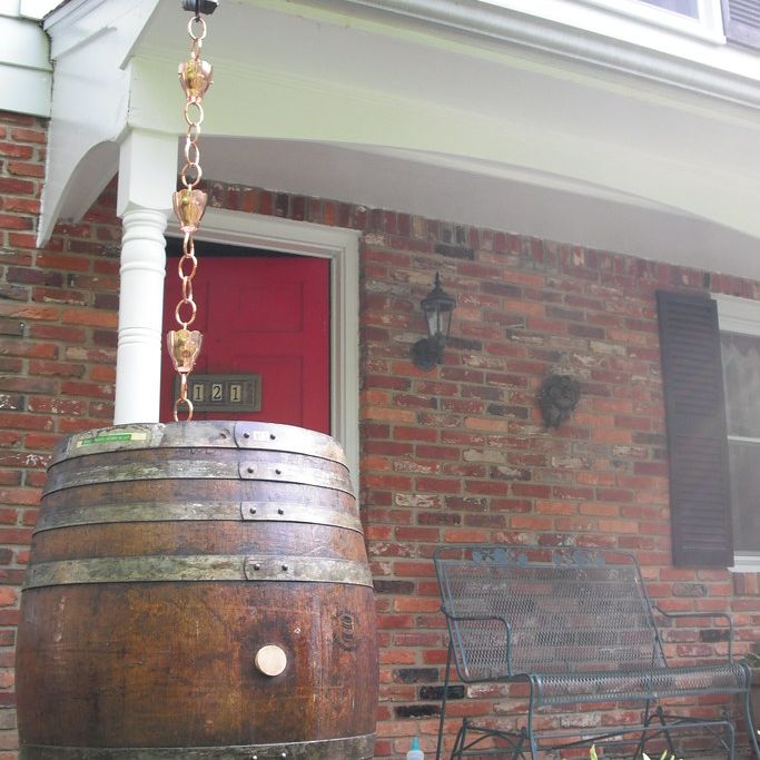 A copper rain chain running from a porch roof into a wooden barrel beside a brick house