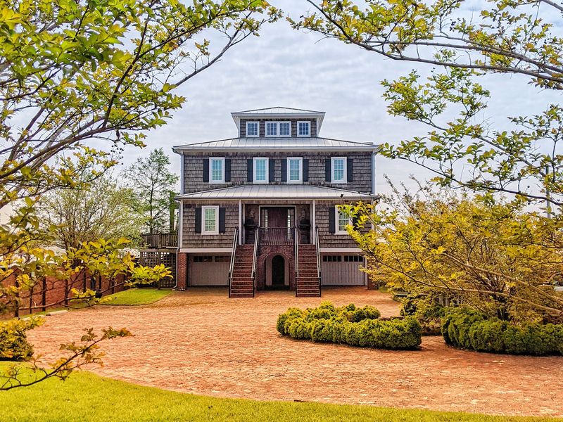 A raised coastal-style house with a metal roof, shingle siding and twin front stairs behind a brick drive