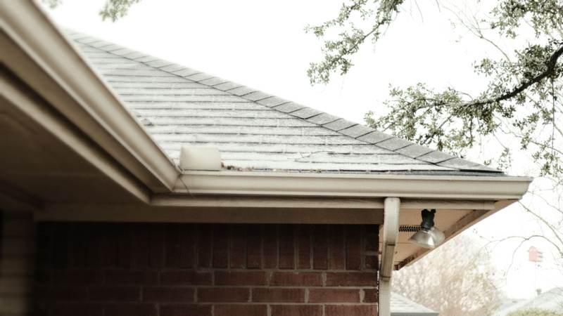 The corner of a red brick house with a white gutter and downspout under overhanging oak branches