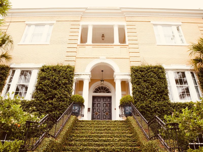 A pale brick two-story home with an arched front door, reached by a wide stair between clipped hedges and iron railings