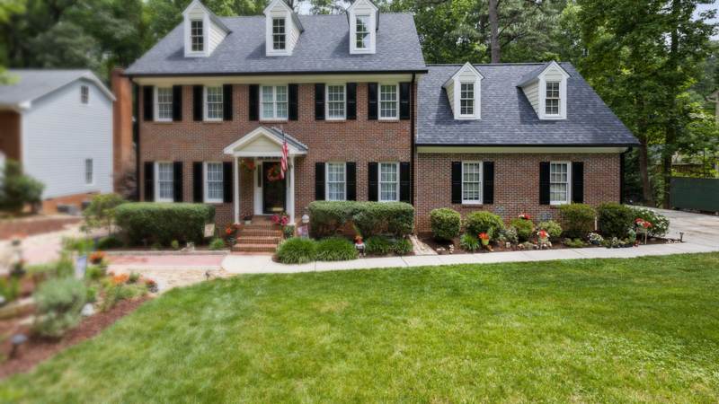 A two-story red brick colonial house with three dormers, black shutters and a flower-lined front walk under tall trees