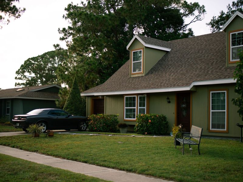 A green two-story house with dormer windows and a lawn, shaded by tall trees