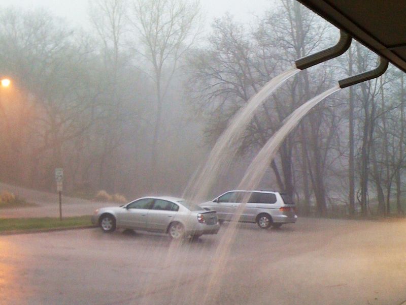 Two downspout outlets shooting arcs of rainwater onto a parking lot during a heavy rainstorm