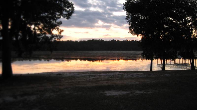 Sunset reflected in still water at the Little Ocmulgee, with silhouetted trees, a pier, a pale sandy shore and a line of pines on the far bank